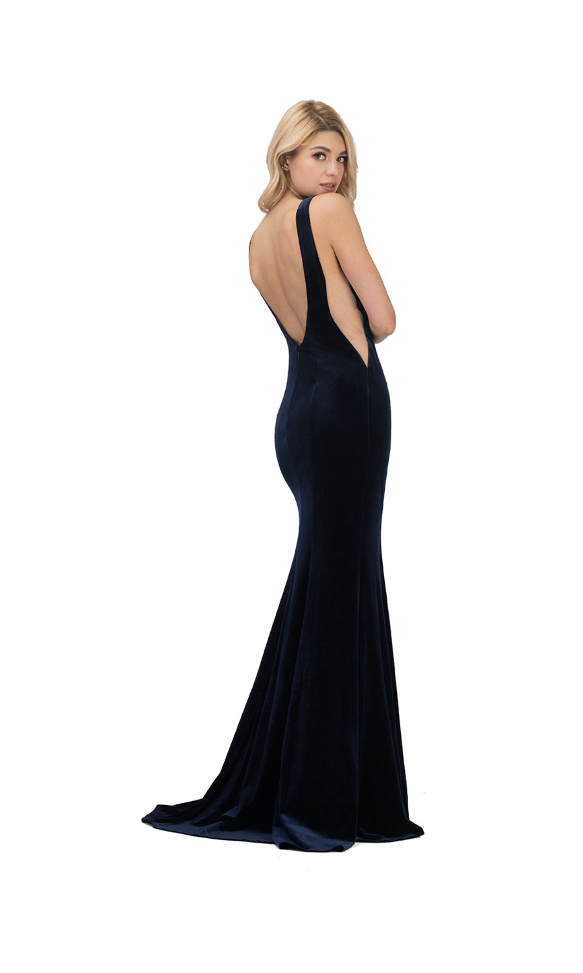 A woman models the Sheer-Sides Long Velvet Prom Dress: Chicas C7052—a fitted, floor-length navy blue gown with an open back—standing against a white background.